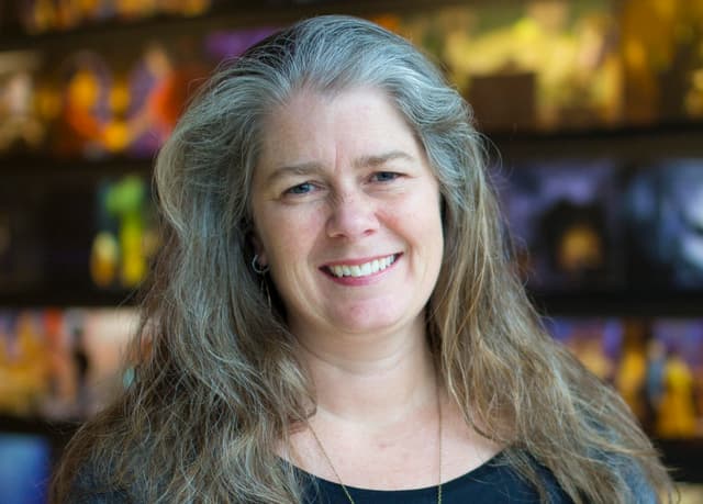 The image shows a smiling woman with long, gray hair against a colorful, blurred background.