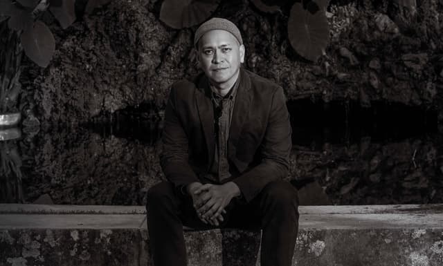 A man with a serious expression sits on a bench in front of a rocky, cave-like background.