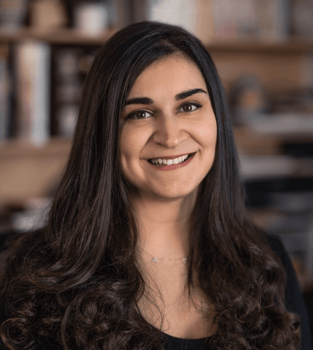 A smiling young woman with long, dark curly hair sits in front of a bookshelf in a cozy, warm-toned setting.