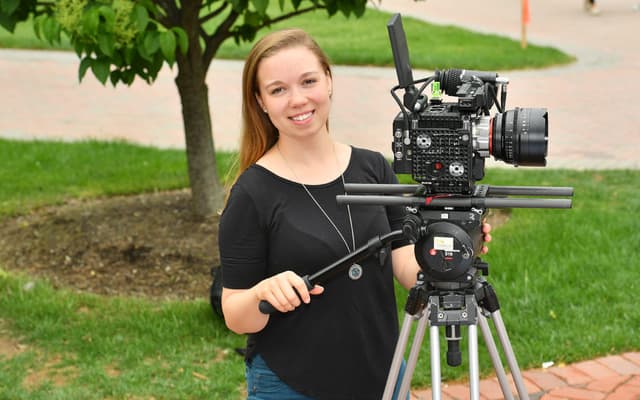A smiling woman in a black shirt stands behind a professional video camera, surrounded by a lush, green outdoor setting.