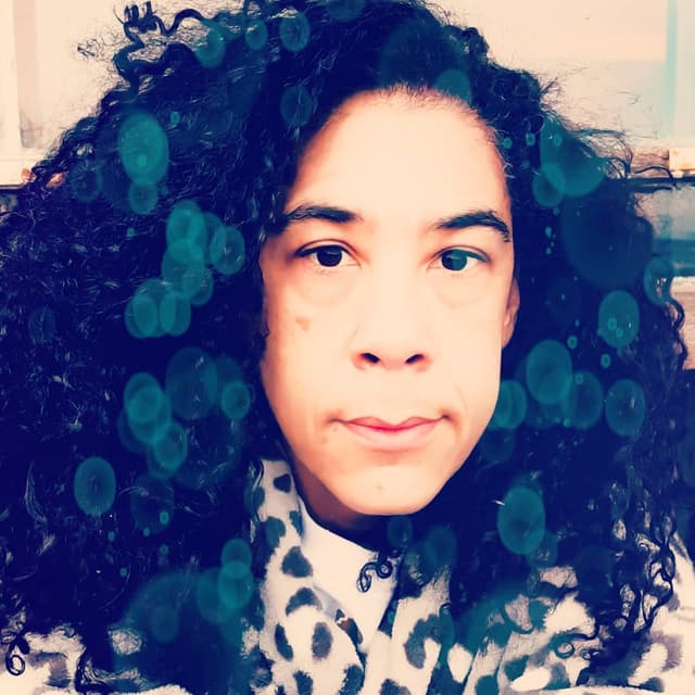 A person with curly dark hair and a patterned scarf appears in the foreground, with a blurred background of what seems to be a room or indoor setting.