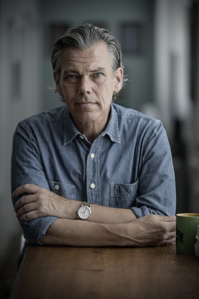 A middle-aged man with gray hair and a serious expression sits at a wooden table, his arms crossed, with a mug in the background.