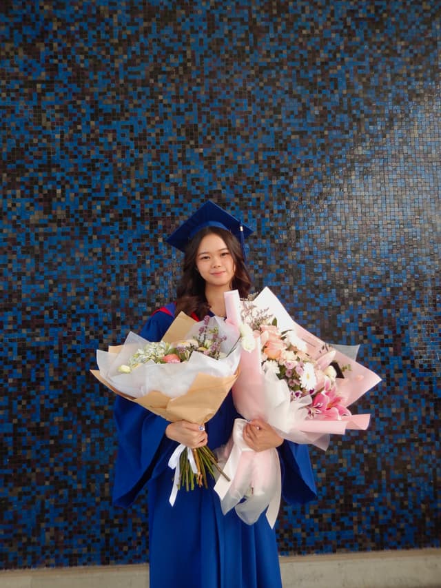 A young woman in a blue graduation gown holds a bouquet of flowers, standing in front of a vibrant blue mosaic wall.