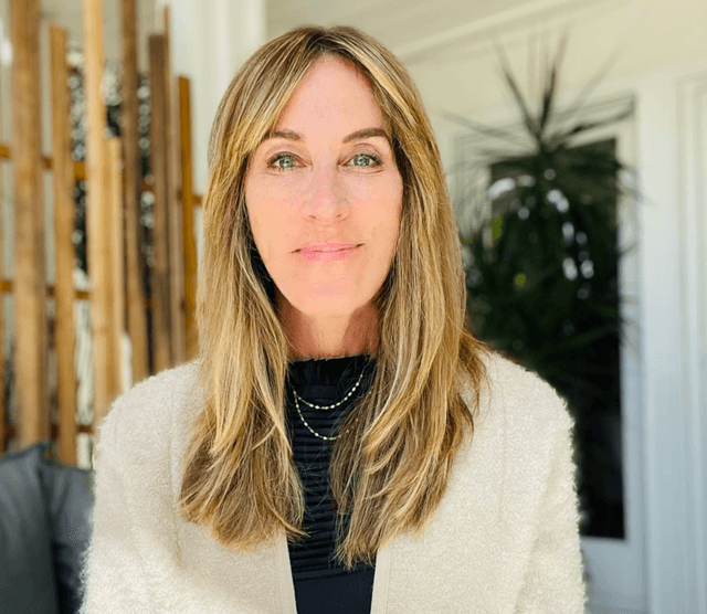 A woman with long, blonde hair wearing a white fuzzy coat stands in front of a wooden structure with plants visible in the background.