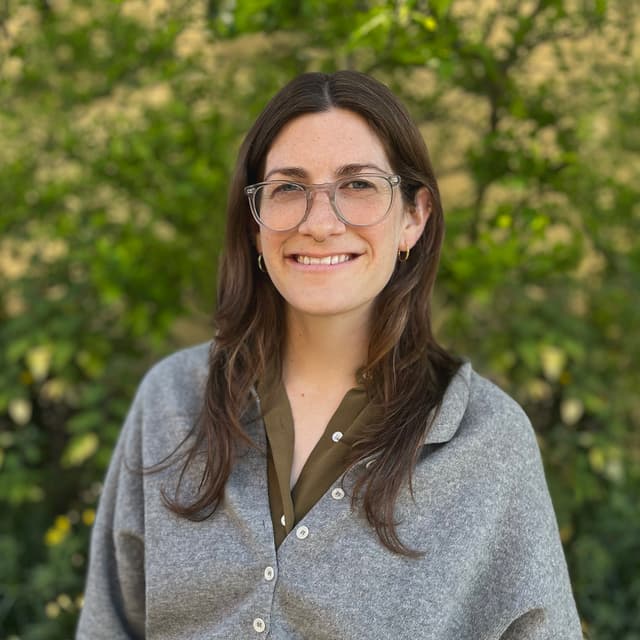 A young woman with long dark hair and glasses is smiling warmly in a natural setting with lush greenery in the background.
