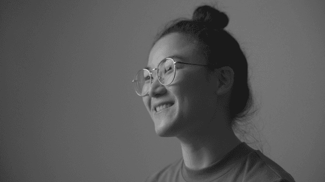 A close-up black and white portrait of a smiling young woman wearing glasses, with her hair tied up in a bun.
