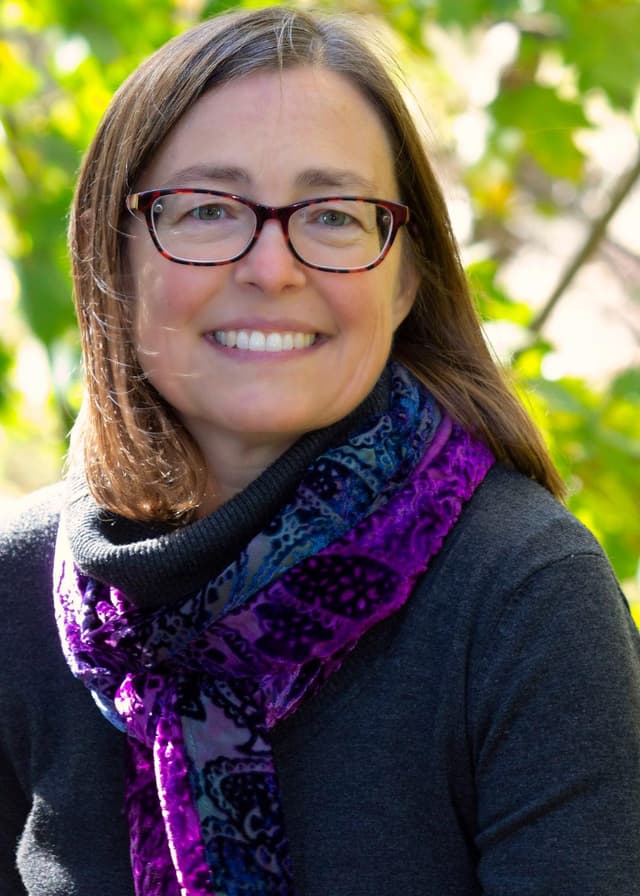 A smiling woman with long brown hair wearing glasses and a colorful scarf stands in a lush, green outdoor setting.
