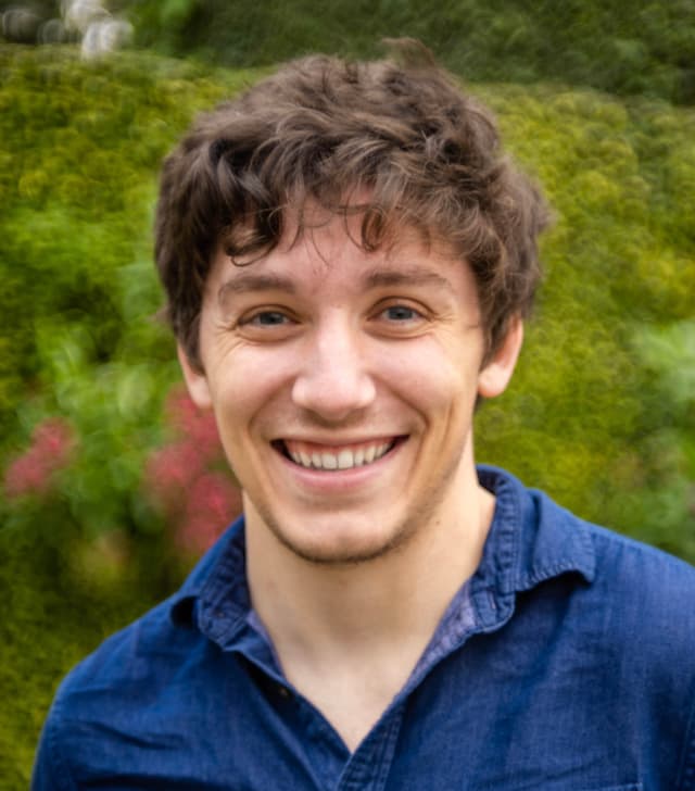 A young man with curly hair and a warm smile stands in a lush, green garden setting.