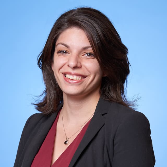 A smiling woman with dark hair wearing a black blazer and red top stands against a bright blue background.