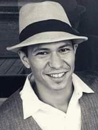 A smiling man wearing a white hat and a light-colored shirt stands in front of a dark background.