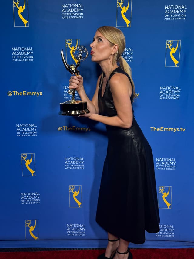 A woman in a black dress is holding an Emmy award trophy while posing in front of a backdrop featuring the National Academy of Television Arts & Sciences logo.