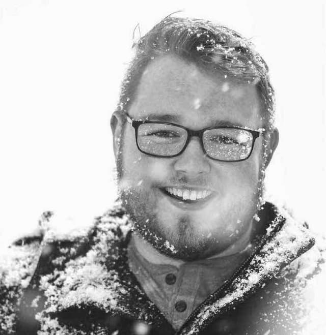 A smiling man with glasses and a beard, surrounded by falling snow, appears in the foreground of the black and white image.
