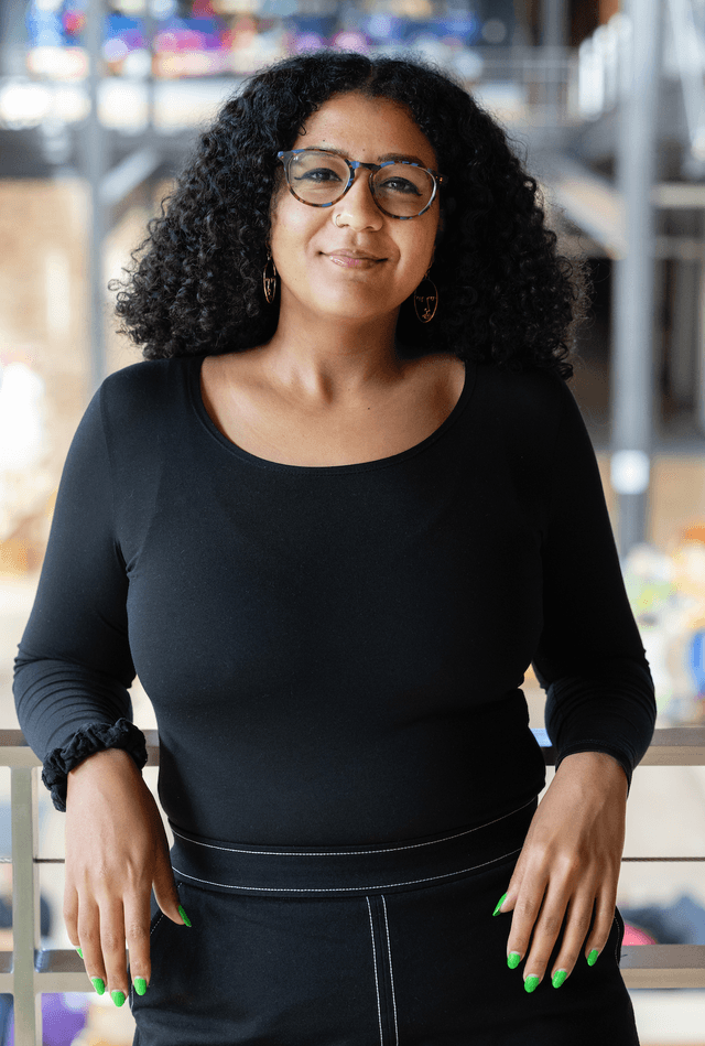 A young woman with curly hair and glasses is smiling while standing in front of a blurred background with colorful lights.