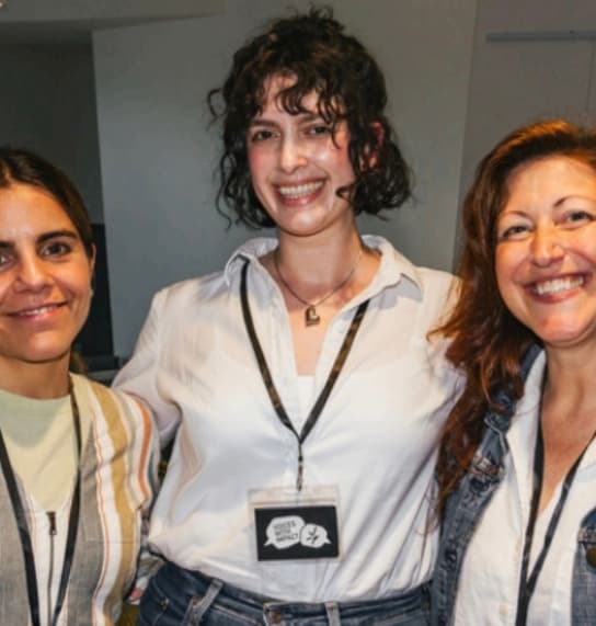 The image shows three smiling women posing together in what appears to be an office or professional setting, with a plain background behind them.