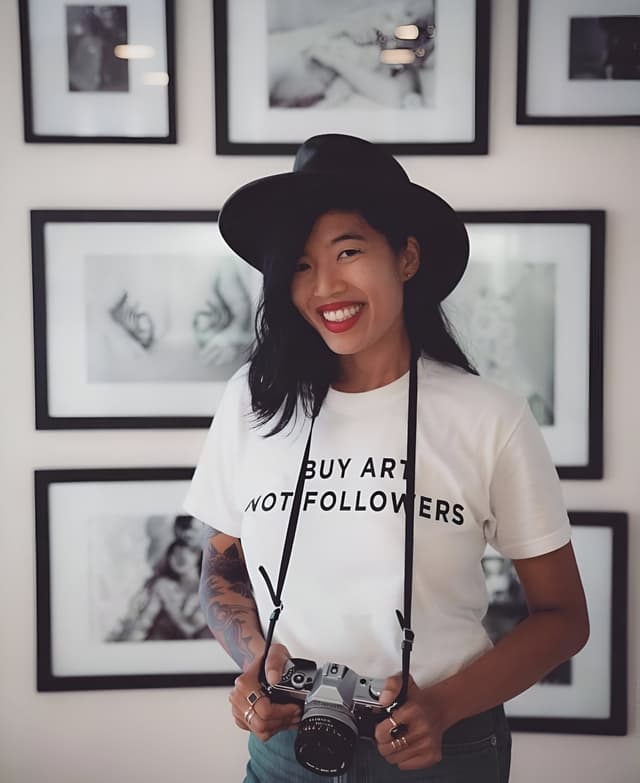 A smiling young woman wearing a white t-shirt with the text "BUY ART NOT FOLLOWERS" stands in front of a wall displaying framed black and white photographs.