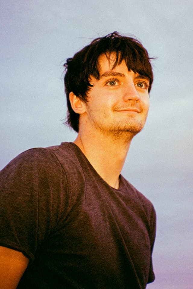 A young man with dark hair and a friendly expression stands against a backdrop of a serene sky at sunset.