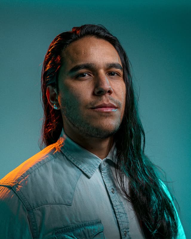 A man with long dark hair and a beard, wearing a gray shirt, stands against a teal background.
