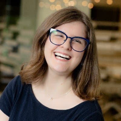 A young woman with long brown hair and glasses is smiling brightly in a cozy indoor setting with warm lighting and decorative elements in the background.