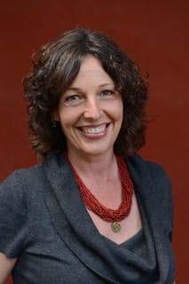 A smiling woman with curly dark hair wearing a red necklace and a dark jacket stands in front of a reddish-brown background.