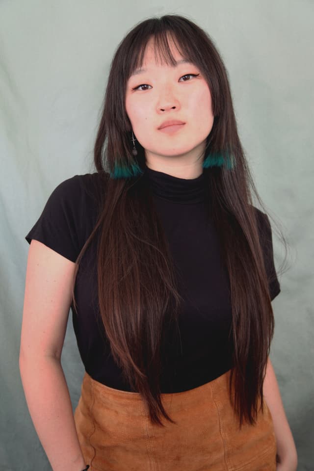 A young woman with long dark hair wearing a black top and a tan skirt stands against a light blue background.