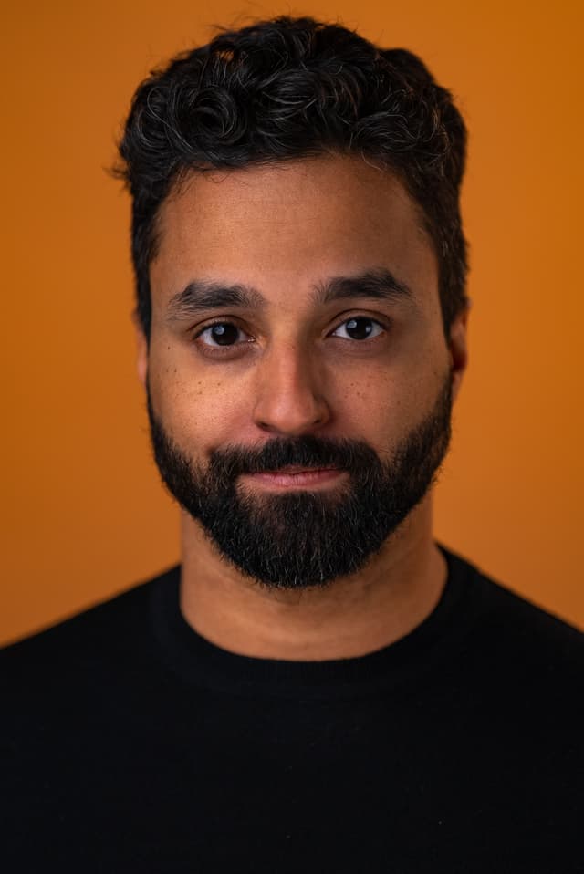 A man with a beard and curly hair is shown against an orange background, looking directly at the camera with a friendly expression.