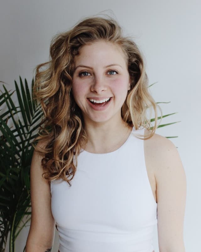 A young woman with curly blonde hair and a bright smile stands in front of a backdrop of lush green plants.