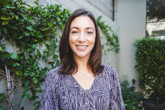 A smiling woman with long brown hair stands in front of a lush, green plant-filled background.