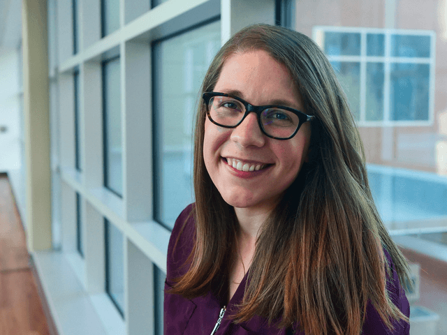 A smiling young woman with long brown hair and glasses stands in front of a window, with a cheerful expression on her face.