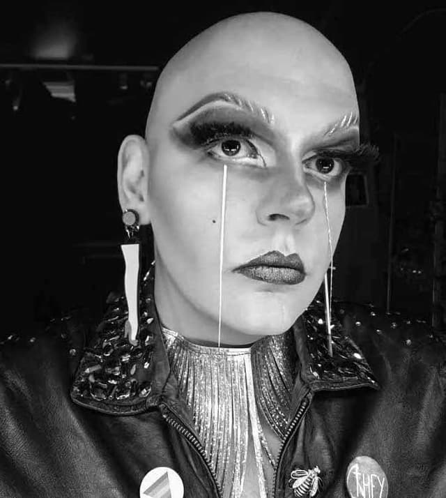 A close-up portrait of a bald individual wearing a metallic, embellished jacket and dramatic makeup, set against a dark background.