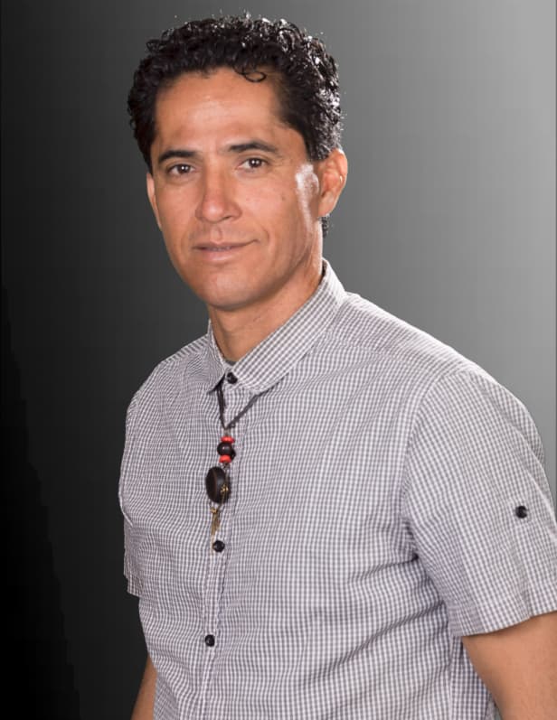 A man with curly dark hair and a friendly expression is wearing a checkered shirt against a plain gray background.